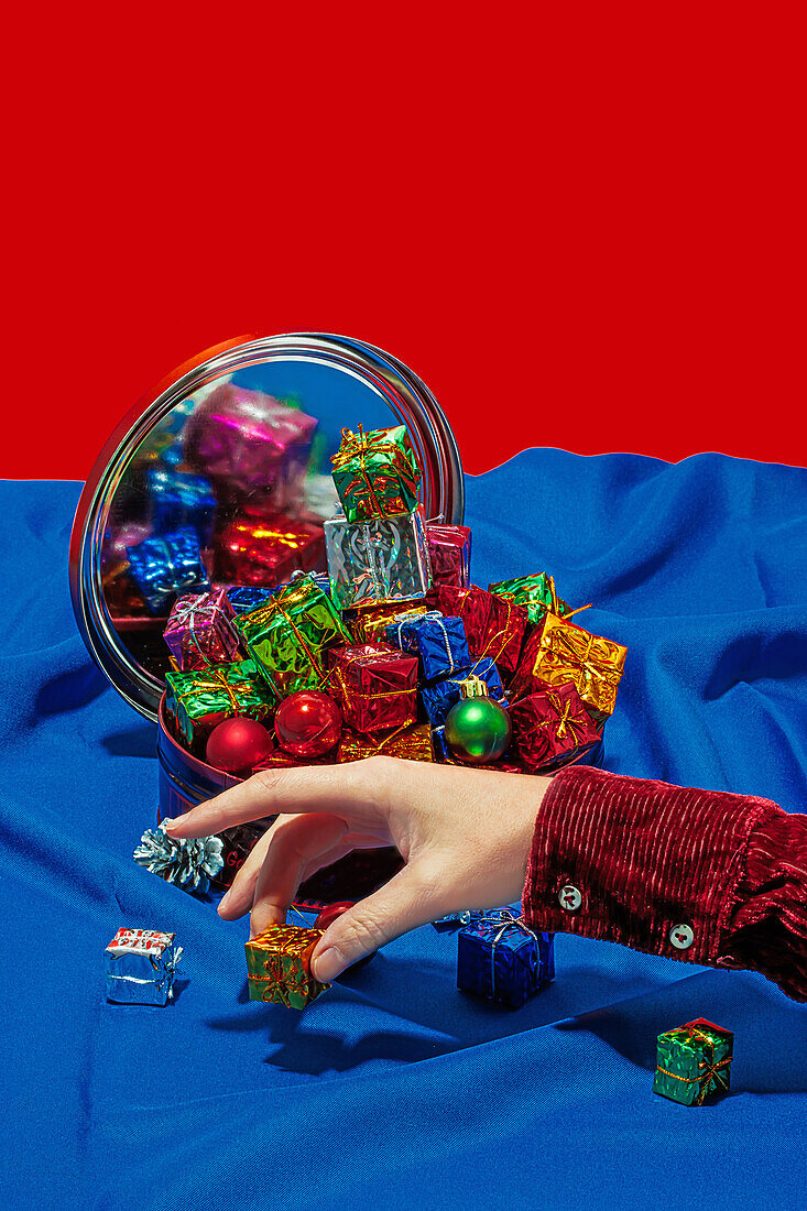 Anonyme Hand in einem festlichen Ärmel greift nach bunten Weihnachtssüßigkeiten, die sich in einem Spiegel auf einem blauen Tischtuch spiegeln