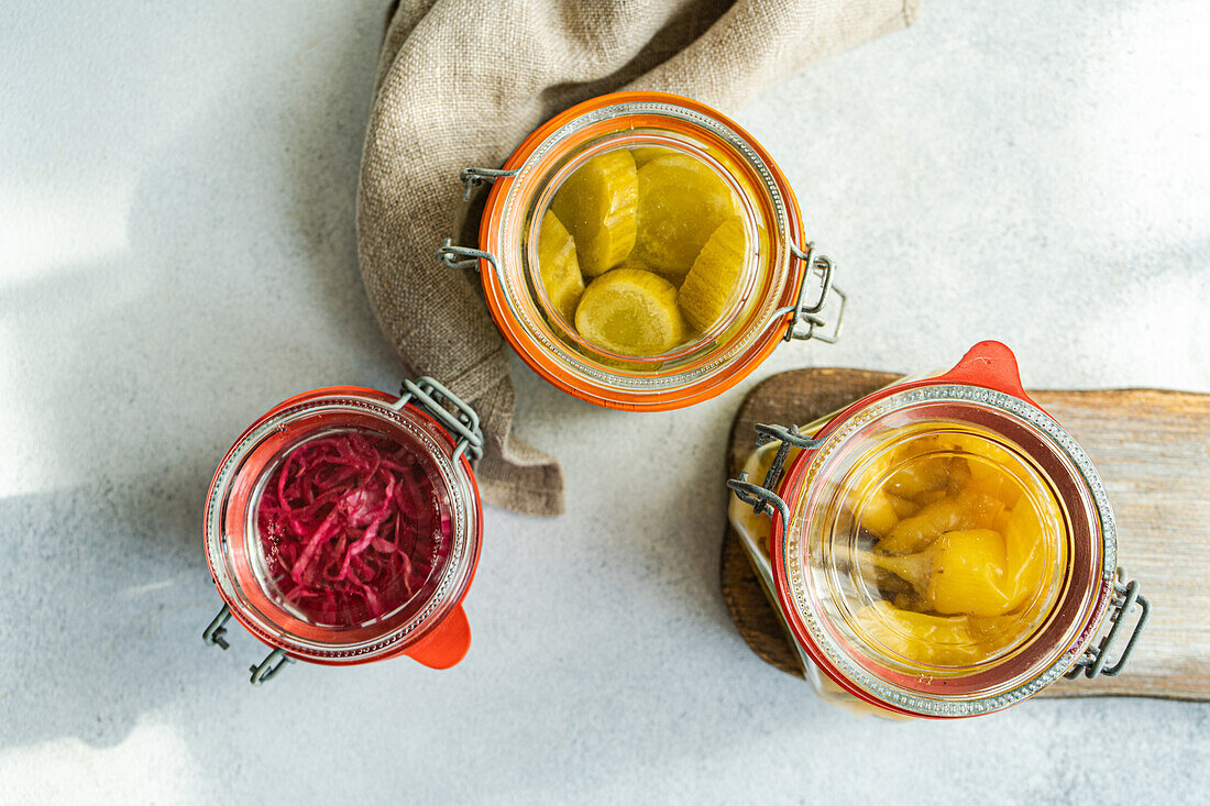 Three glass jars filled with colorful fermented vegetables, including pink cabbage with beetroot, yellow white cucumbers, and hot spicy peppers, placed on a wooden board with a neutral background.