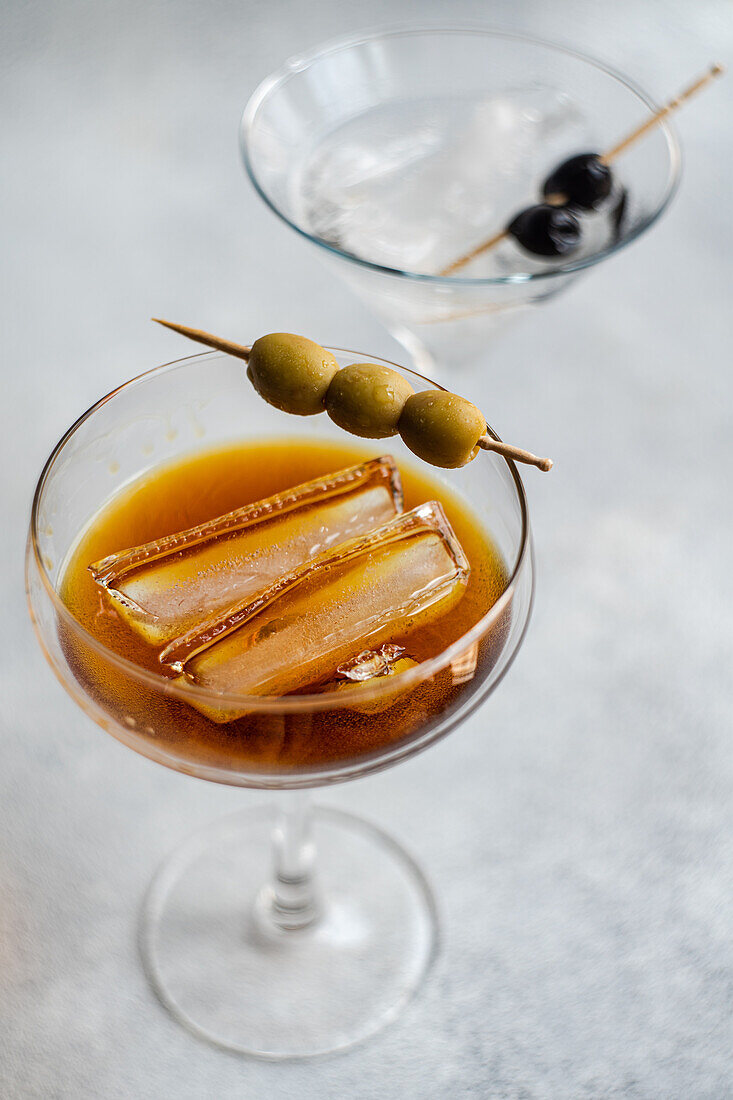 High angle of glasses of alcoholic cocktails with whiskey and sticks of oily olives slices on gray background in studio