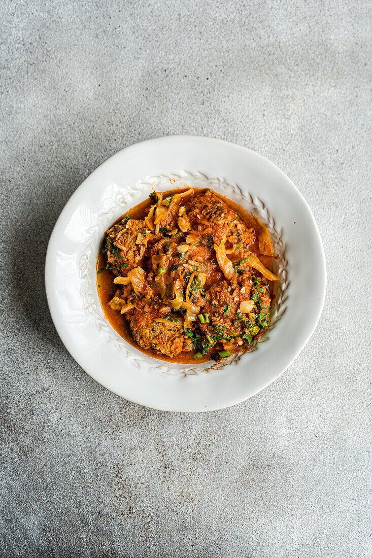 Draufsicht auf ein georgisches Gupta-Gericht mit Fleischbällchen und Reis, geschmort in Tomatensoße mit Kohl, Karotten und Zwiebeln, serviert mit gehackten Koriandern auf einem Teller neben Löffel, Gabel und Serviette vor grauem Hintergrund
