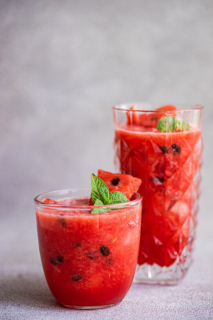 Glasses of Margarita cocktail with watermelon smoothie garnished with leaves of mint placed on gray table
