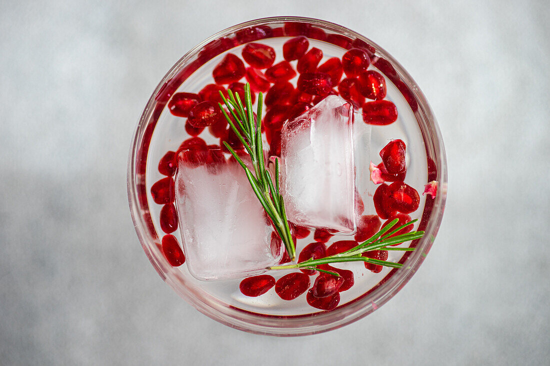 From above of gin tonic cocktail adorned with pomegranate seeds and rosemary in a wide-brimmed glass on a textured grey background