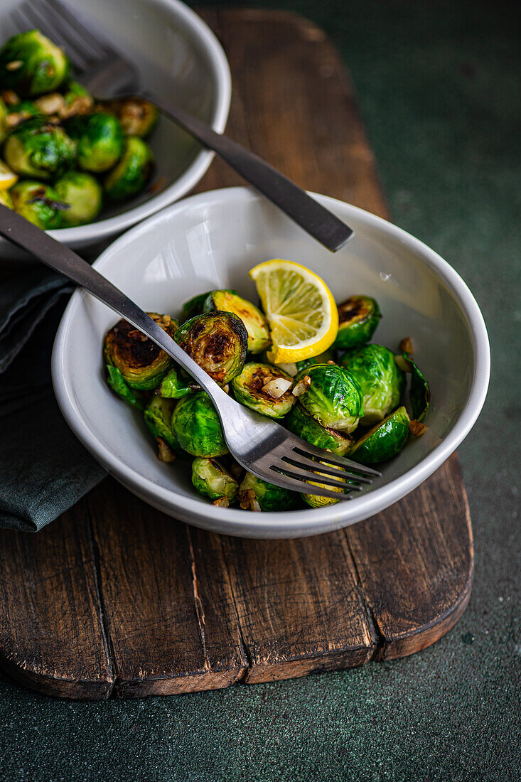 From above of bowl of grilled Brussels sprouts seasoned with garlic and spices, garnished with a slice of lemon, served on a wooden board with a fork