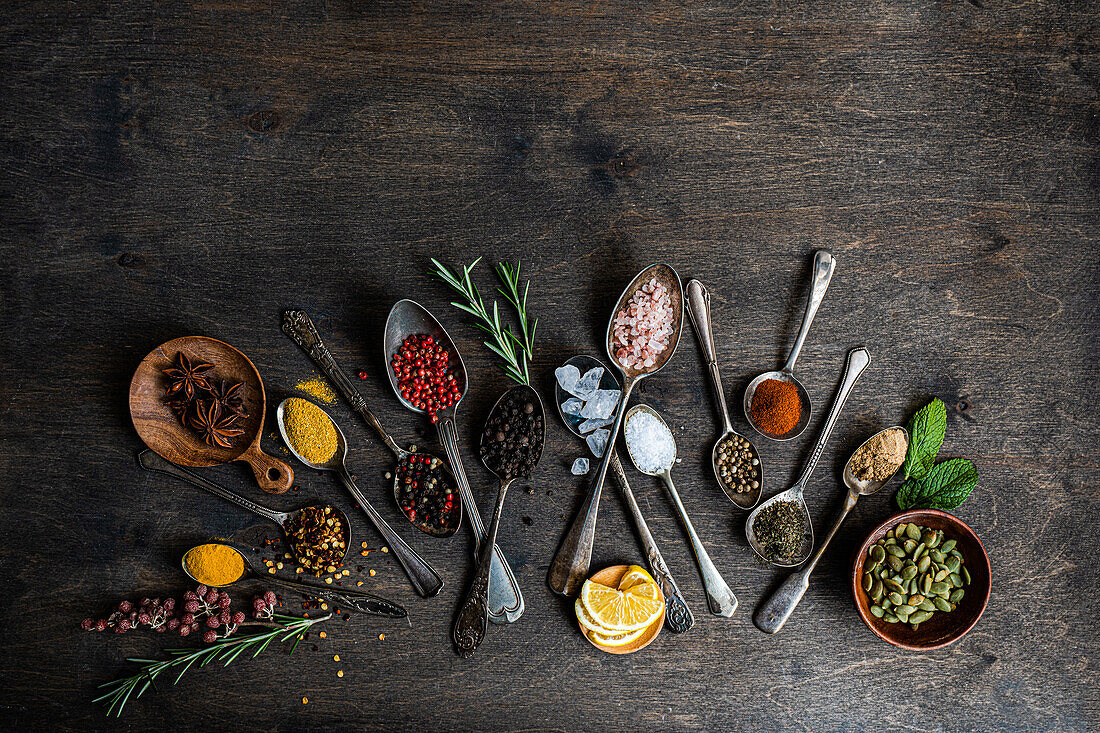 From above flat lay showcasing a variety of spices and seeds in spoons on a dark wooden surface, including sunflower seeds, anise stars, rosemary, mint, lemon, and various colorful peppers and salts.
