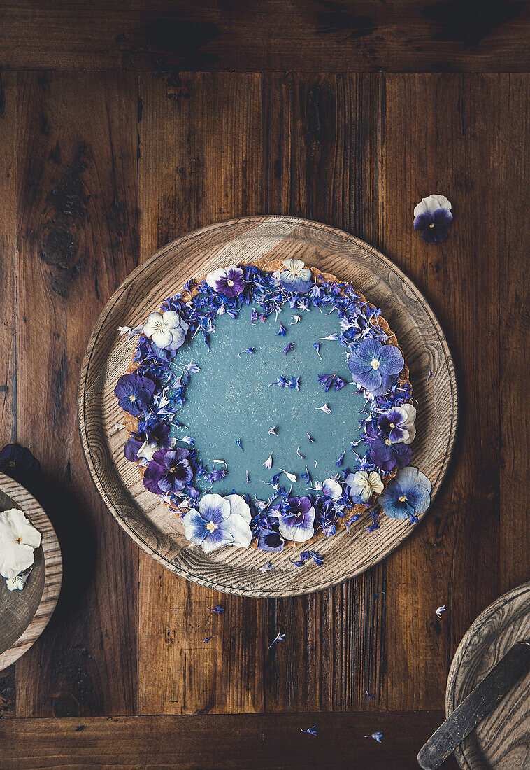 Draufsicht auf eine runde Torte aus blauer Spirulina und mit Blumen dekoriert auf einem Holztisch