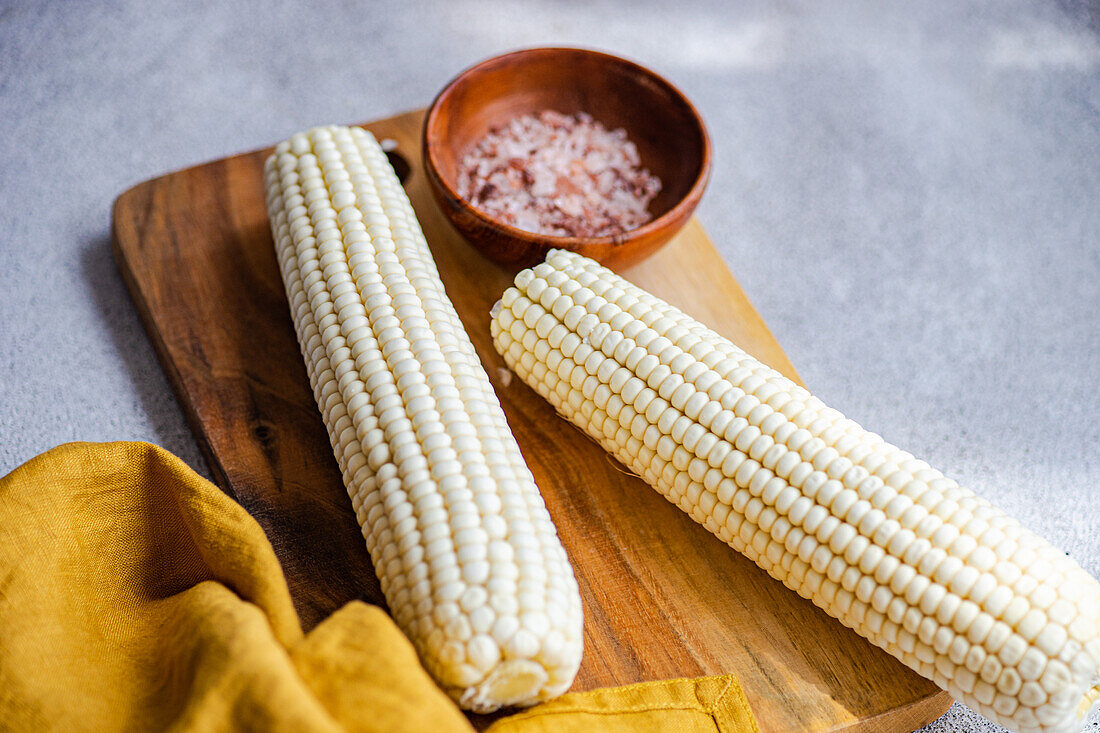 From above wooden chopping board with ripe corncobs placed on concrete table in kitchen with bowl with sea salt and napkin