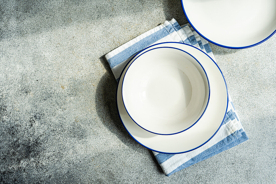 Draufsicht auf einen minimalistisch-rustikal gedeckten Tisch mit weißen Tellern und gestreifter Serviette auf grauer Fläche im Tageslicht