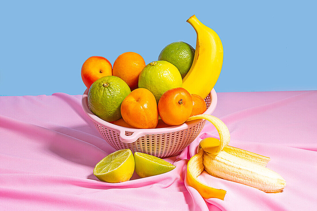 Plastic bowl with fresh assorted fruits placed on at table with a pink tablecloth table in daylight on blue background