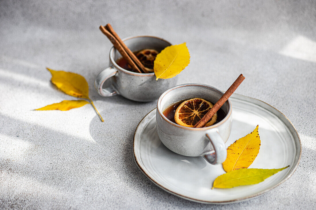 A warm cup of spiced tea garnished with cinnamon sticks, anise and dried orange slices complemented by vibrant yellow autumnal leaves on a light gray surface