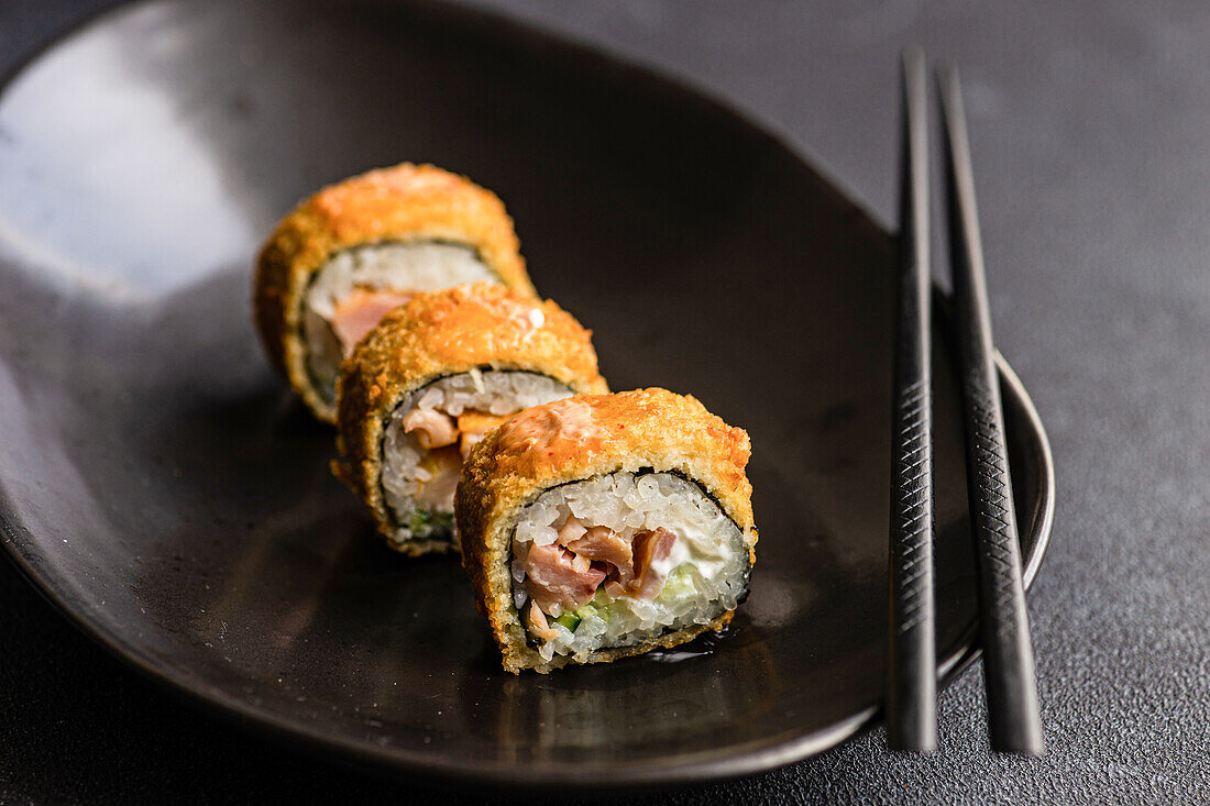 Focused row of tasty fried sushi rolls with cooked rice and salmon with avocado slices on ceramic black plate on table