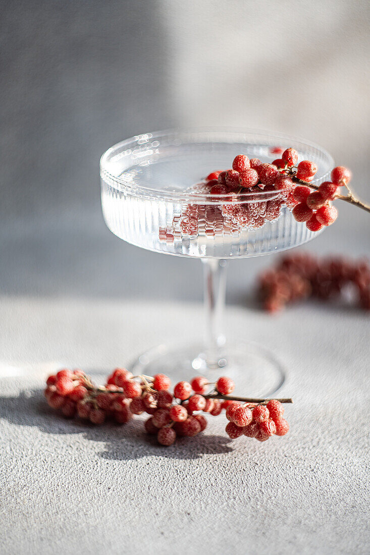A sophisticated cocktail glass filled with a clear beverage, adorned with a sprig of red berries on a textured surface, illuminated by natural light