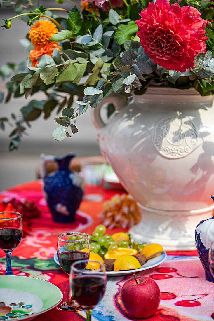 A colorful table setting with wine glasses, fruits on a plate, and a blue ornate vase against a red patterned cloth.