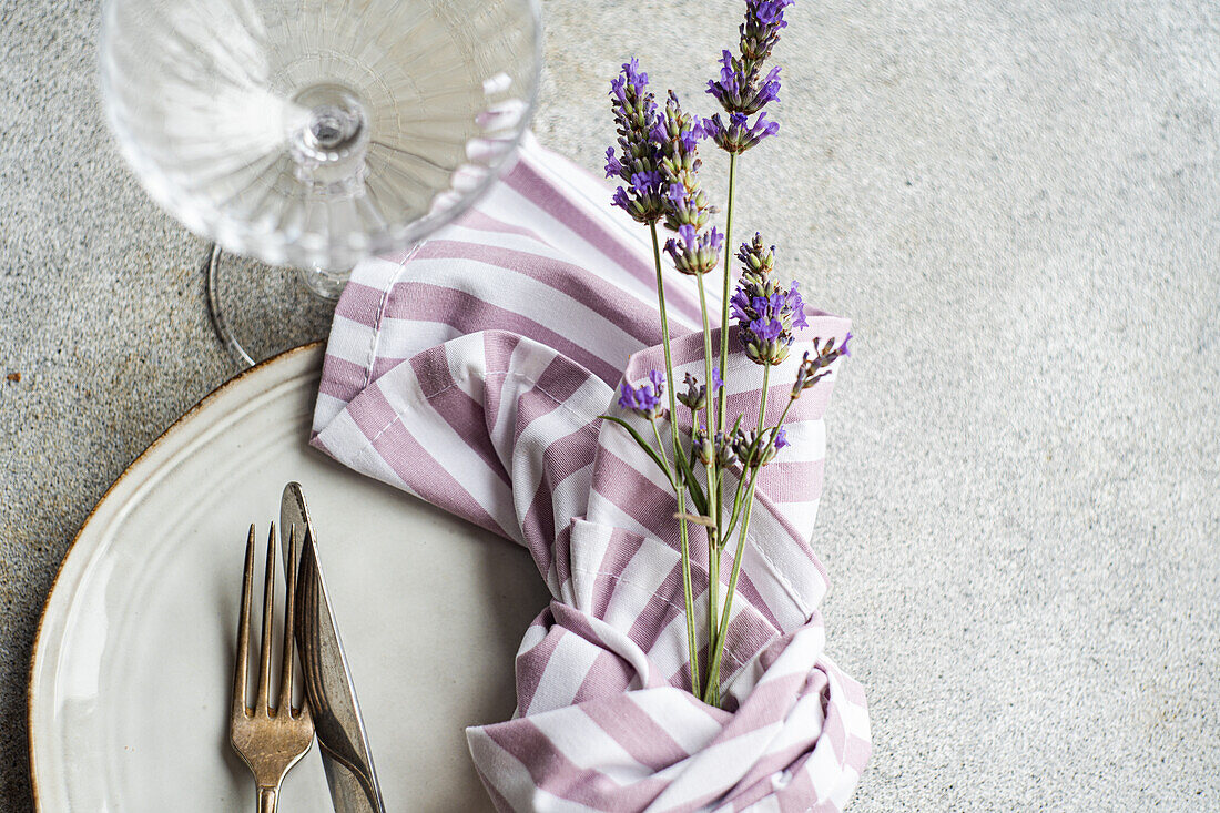 Draufsicht auf arrangierte Blumen in Serviette und Gabel auf weißem Keramikteller mit Messer auf grauem Untergrund