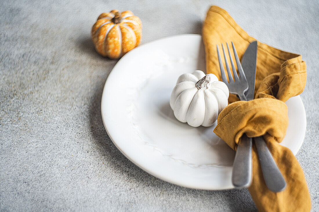 White ceramic plate with decorative pumpkins with silver fork and knife, wrapped in a mustard-colored napkin, placed on a textured gray concrete surface