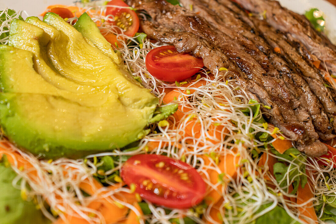 Top view of appetizing grilled beef served on white plate with cut fresh vegetables avocado tomato carrot sprouts green leaves in light