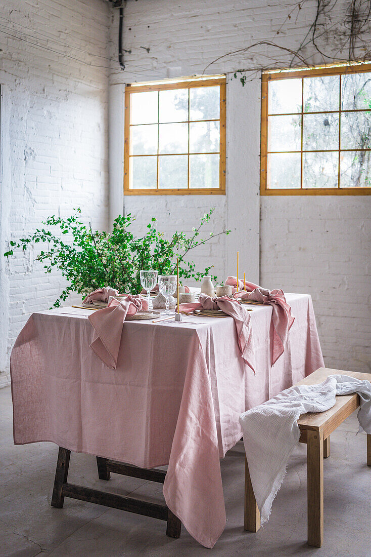 Holztisch mit rosafarbener Tischdecke und Geschirr neben einer mit Stoff bezogenen Bank vor einer Topfpflanze und einer weißen Backsteinmauer