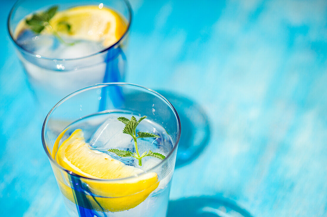Hochformatiger Sommercocktail mit Zitronenwodka, Zitronenscheiben und Minzblättern, serviert mit Eis auf einem blauen Tisch
