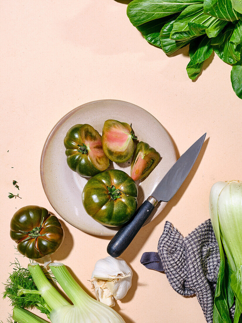 Grüne Tomaten mit anderem Gemüse wie Bok Choy in hellem Sonnenlicht flachgelegter Teller mit Messer mit Kopierraum auf beigem Hintergrund