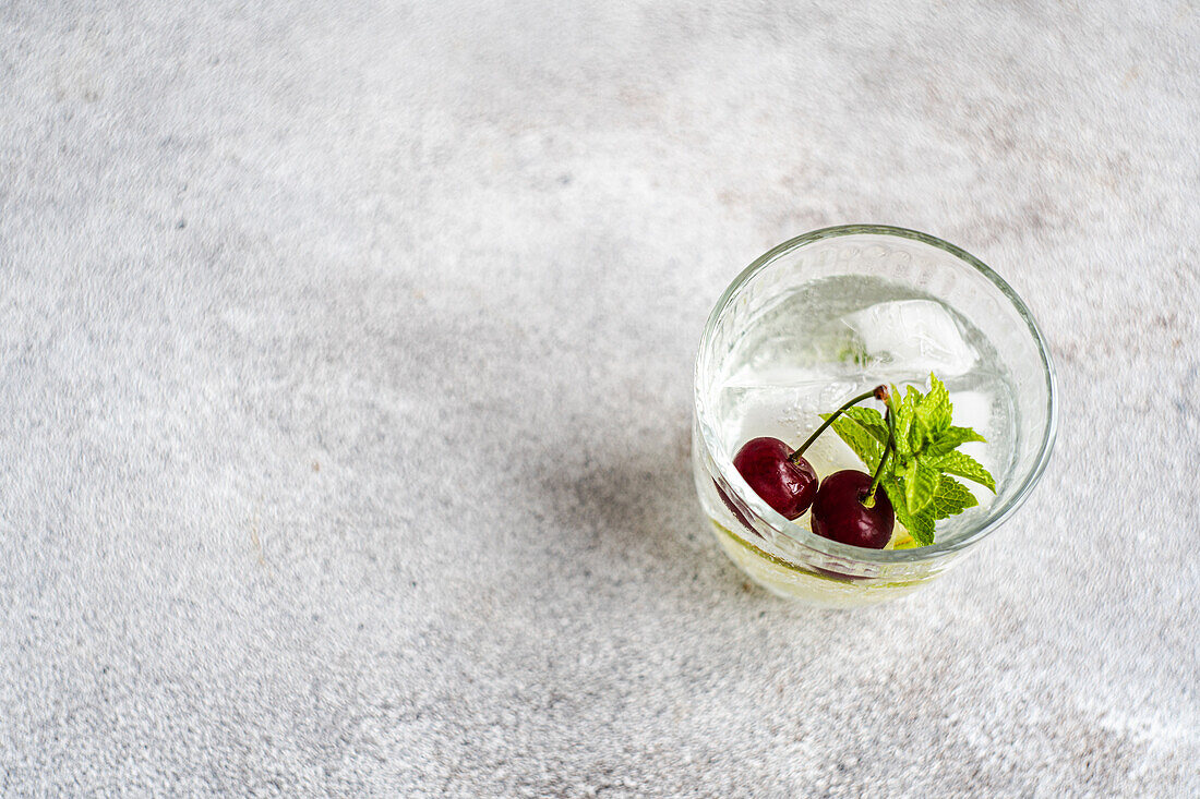 From above transparent glass of refreshing cold drink with lime and cherry and mint leaves on gray surface in summer day against blurred background
