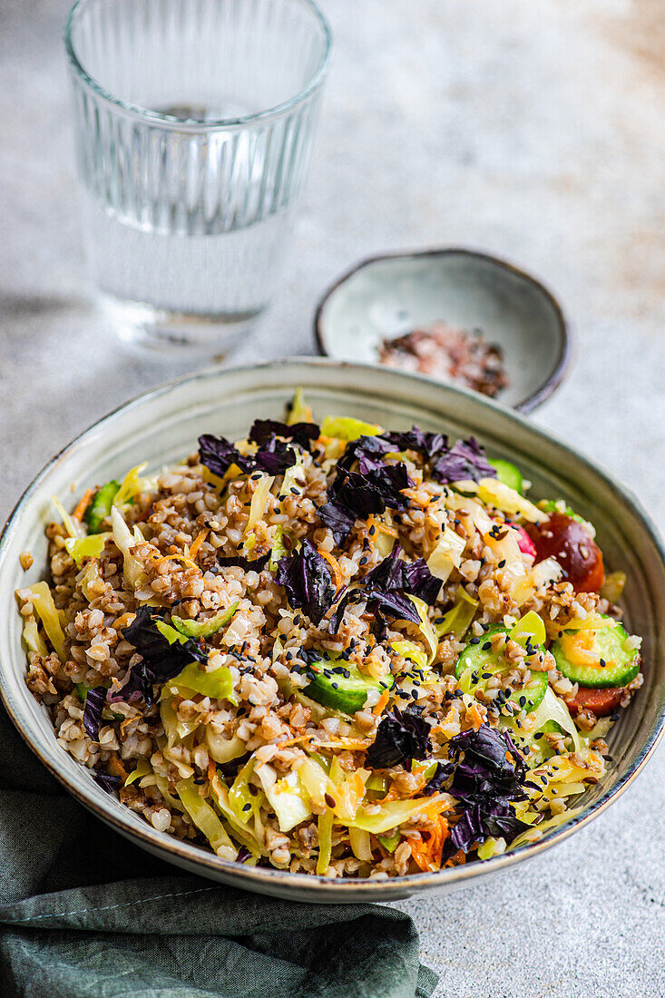 High angle of bowl with delicious buckwheat containing fresh basil plant with cabbage and cucumber with tomato and onion placed on gray table with cup