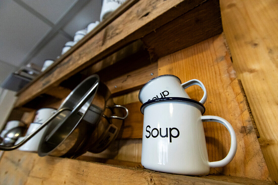 Low angle of white soup mugs stacked on a rustic wooden kitchen shelf, with metal pots hanging below, creating a cozy and organized culinary setting