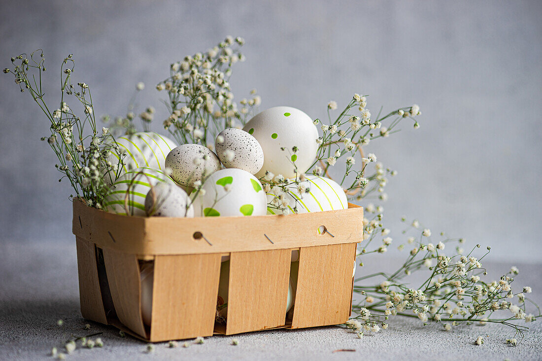 A wooden basket filled with patterned Easter eggs, adorned with delicate gypsophila flowers on a textured background
