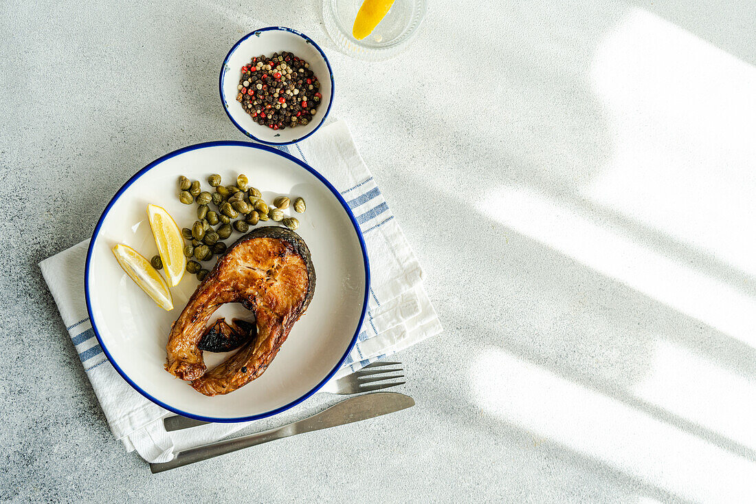 Draufsicht auf ein gut zubereitetes gegrilltes Forellensteak mit Kapern und Zitrone, serviert auf einem weißen Teller mit blauem Rand, darauf eine gestreifte Serviette, Besteck und eine Schale mit Kapern