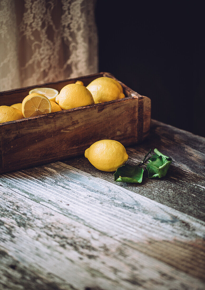 Hoher Winkel von reifen Zitronen und geschnittenen Stücken in einer Holzkiste auf einem Tisch im Tageslicht vor dunklem Hintergrund in einer Küche
