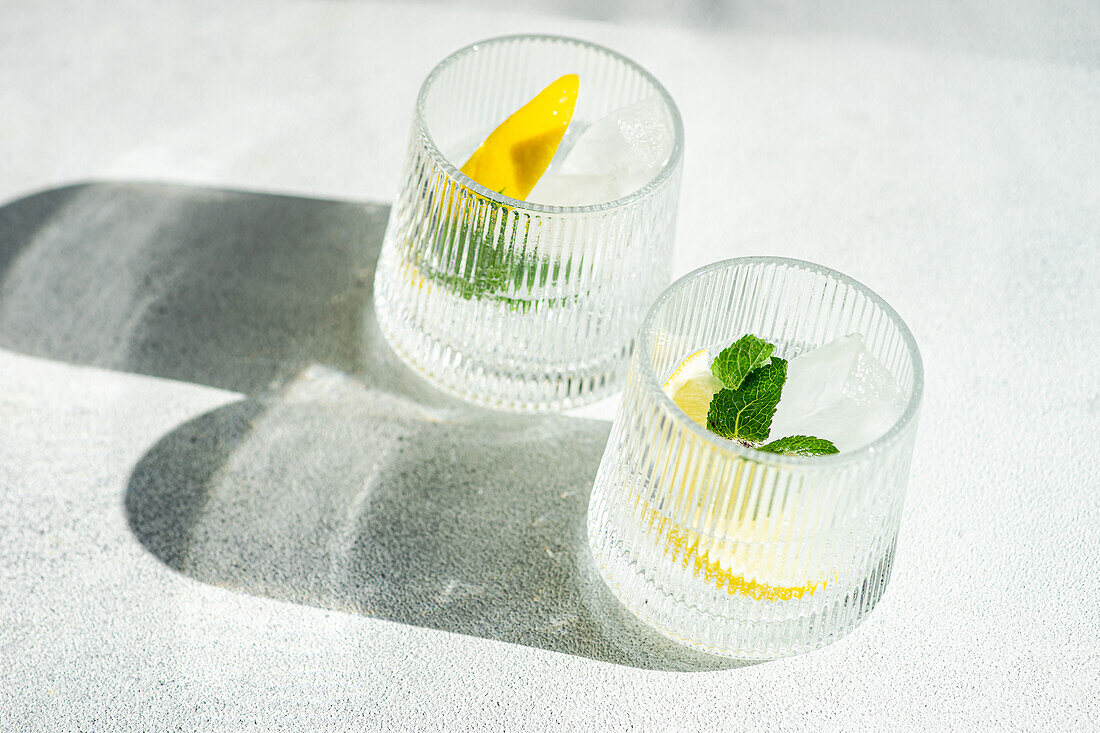 Zwei Gläser mit eiskaltem Gin Tonic, verziert mit Zitrone und Minze, auf einer sonnigen Fläche, die weiche Schatten wirft