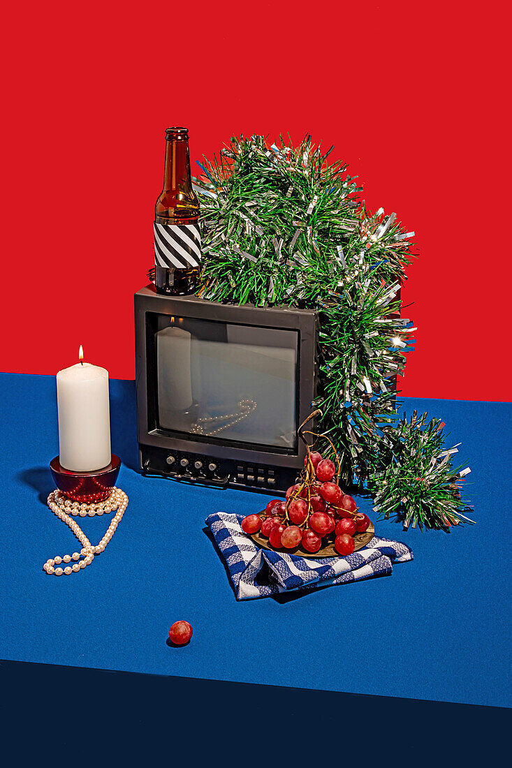 High angle of vintage television set surrounded by an array of objects, including a bottle with a striped label, fresh grapes on a checkered cloth, a white candle and green tinsel, all set against a red backdrop on a blue table