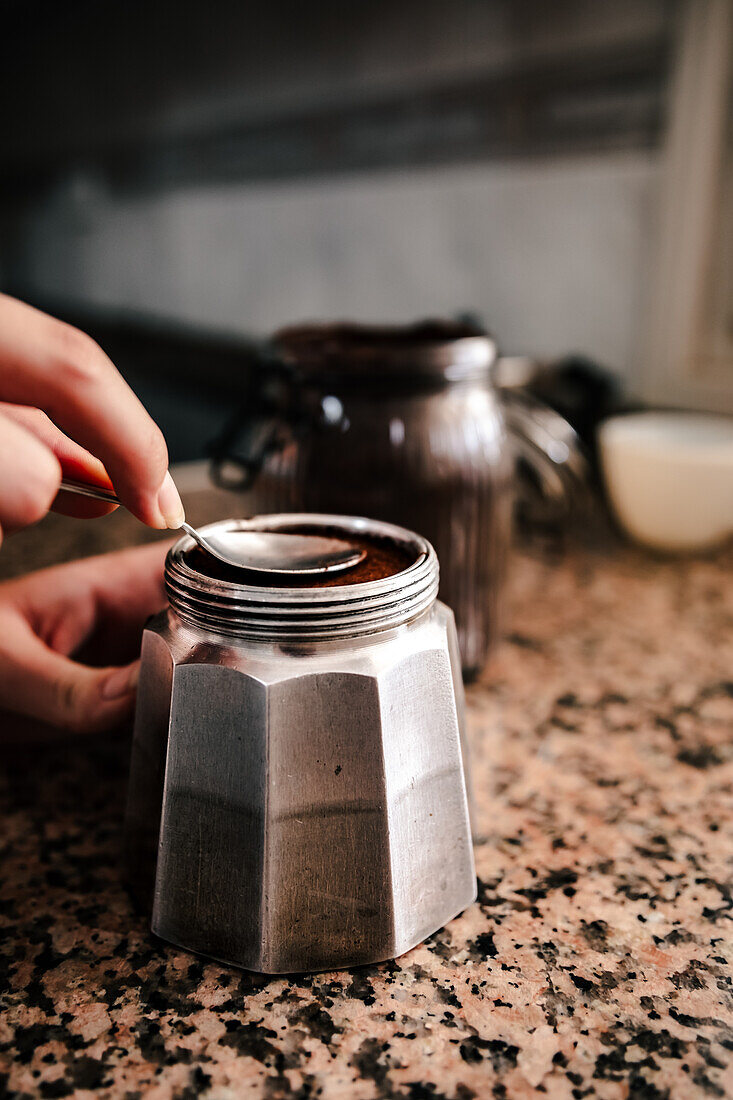 Close-up of anonymous young individual's hand using a spoon to open or seal a stainless steel stovetop espresso maker, amidst a home kitchen setting with a coffee jar in the backdrop