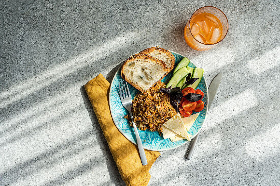 Draufsicht auf fünf Zutaten Mittagessen Set besteht aus hausgemachten Ziegenkäse, frische Gurken und Tomaten mit Blättern von Basilikum Kraut, zusätzlich zu gebratenem Reis mit Fleisch mit Scheiben von gesunden Sauerteig Brot serviert mit Glas Karotte-Apfel-Saft gegen grauen Hintergrund