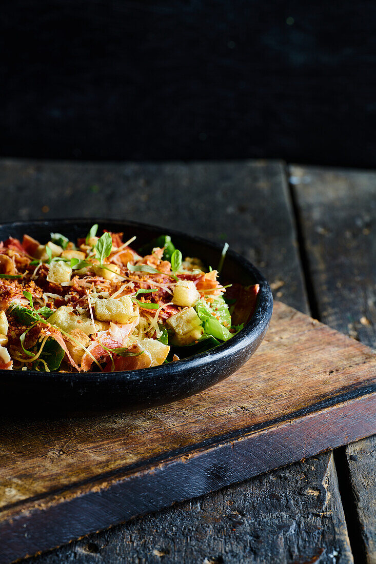 Hoher Winkel von appetitlichen Salat, bestehend aus frischem gehackten Gemüse, Kräutern und Speck in einer schwarzen Schüssel serviert