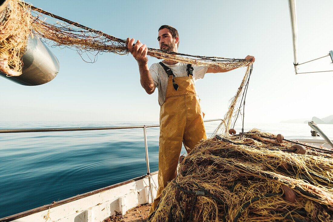 Von unten fokussierter bärtiger männlicher Fischer in Uniform, der mit einem Netz auf Fischfang geht, während er auf einem Schoner in Soller nahe der Baleareninsel Mallorca arbeitet