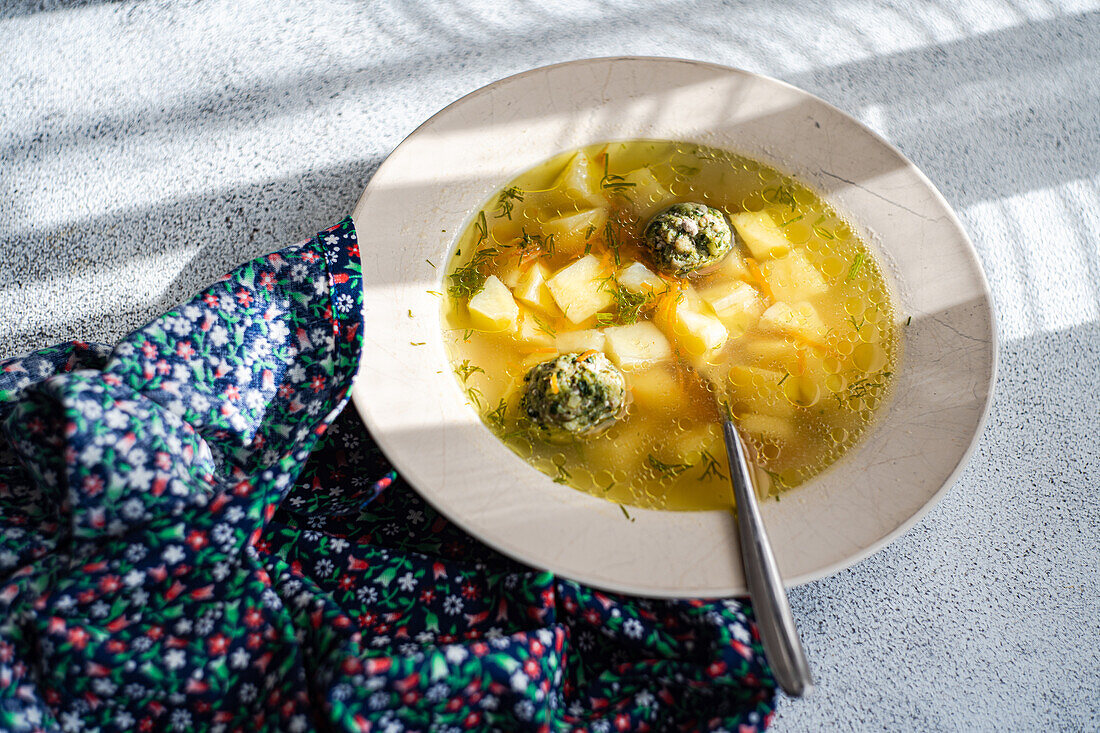 A bowl of meatball soup with potatoes and herbs, shot in natural sunlight, casting shadows.
