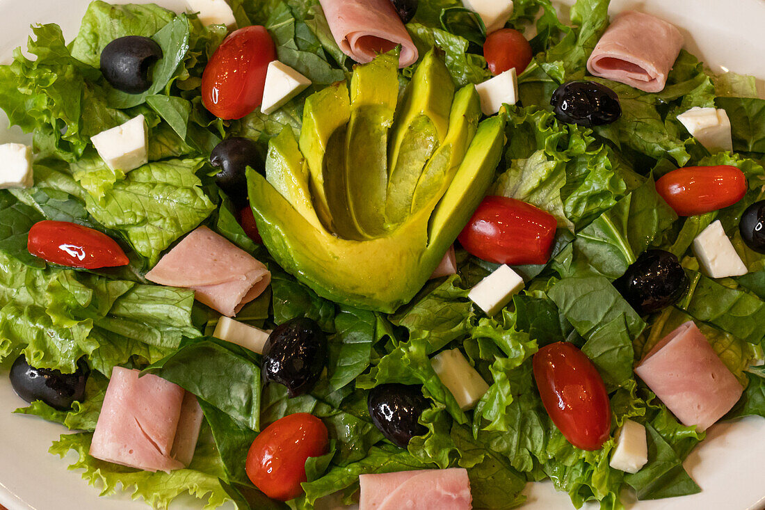 Draufsicht auf einen appetitlichen gemischten Salat mit Avocadostücken, ungeschnittenen Tomaten, schwarzen Oliven, die auf einem geschnittenen grünen Blattsalat liegen, Käse, Salami, Peperoncini und auf einem Teller im Licht serviert