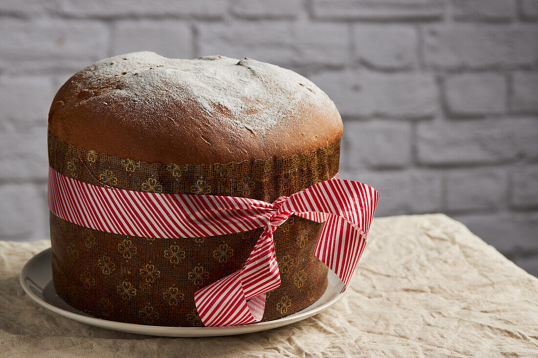 A freshly baked panettone, adorned with a decorative ribbon, stands against a rustic brick background