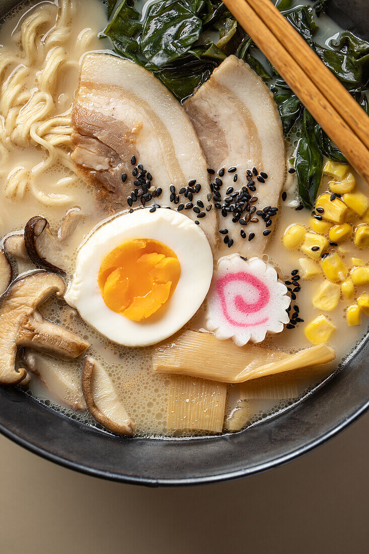 From above of appetizing Japanese ramen with boiled egg and mushrooms served in bowl with wooden chopsticks against beige background