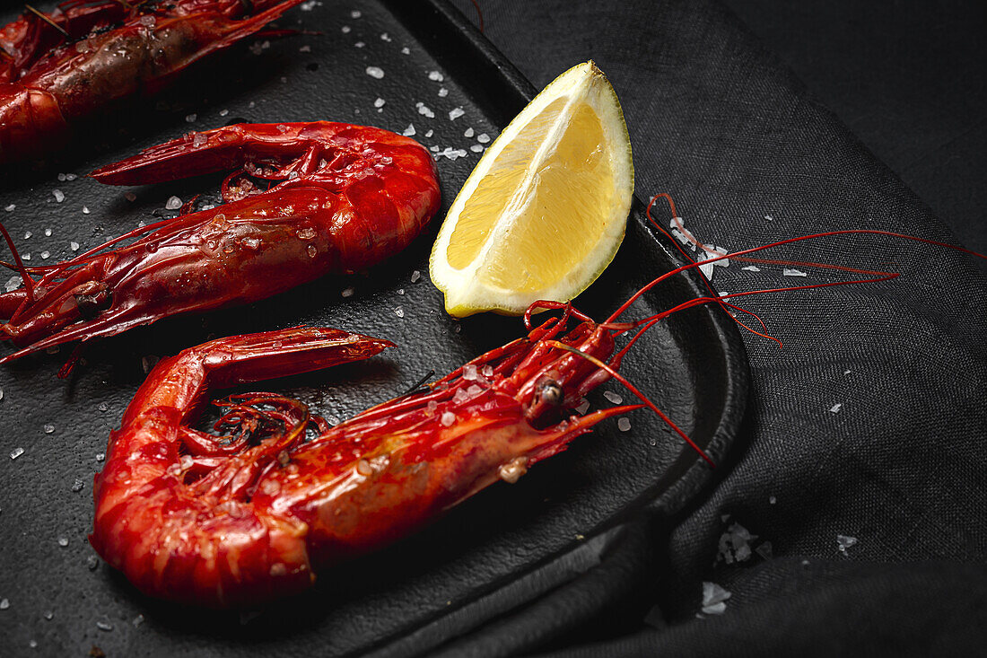 Delicious cooked red prawns on tray with coarse salt and juicy lemon pieces on dark background