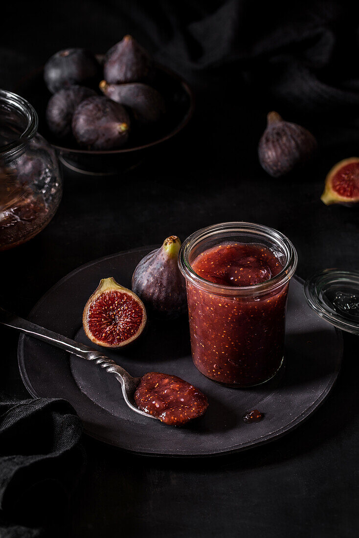From above fresh fruit jam in small jar and on spoon with full and cut figs on plate at black kitchen table