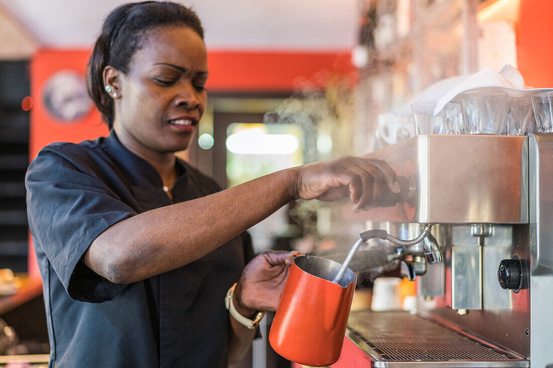 Afroamerikanische Barkeeperin, die Milch in einem Metallkrug aufschlägt, während sie ein köstliches Getränk in einer Kaffeemaschine zubereitet
