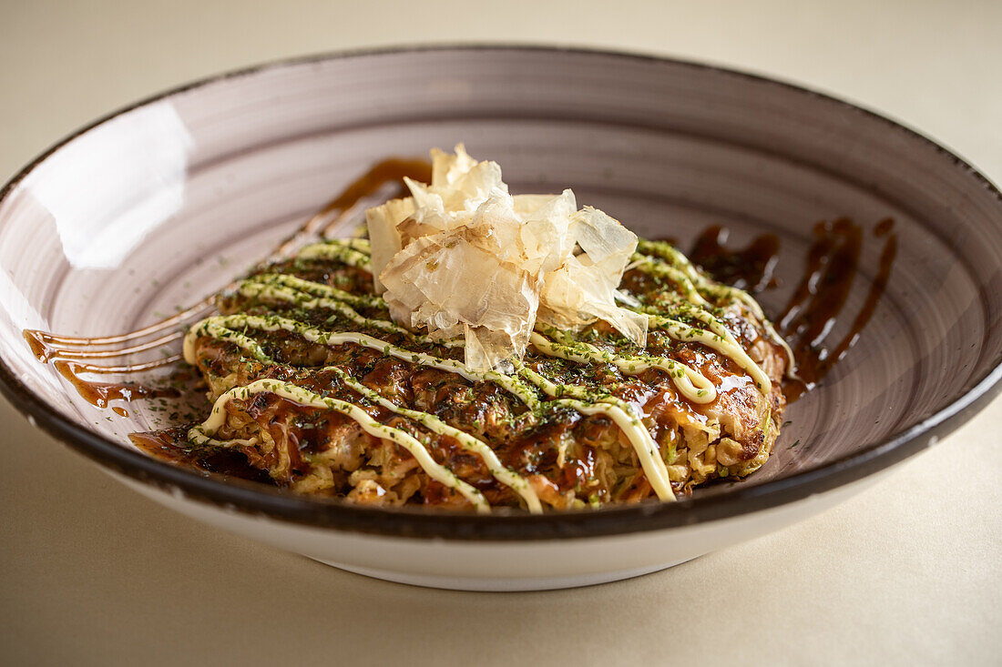 Von oben schmackhaftes traditionelles japanisches Okonomiyaki-Gericht mit Soße und Bonitoflocken in Keramikschüssel auf hellem Hintergrund