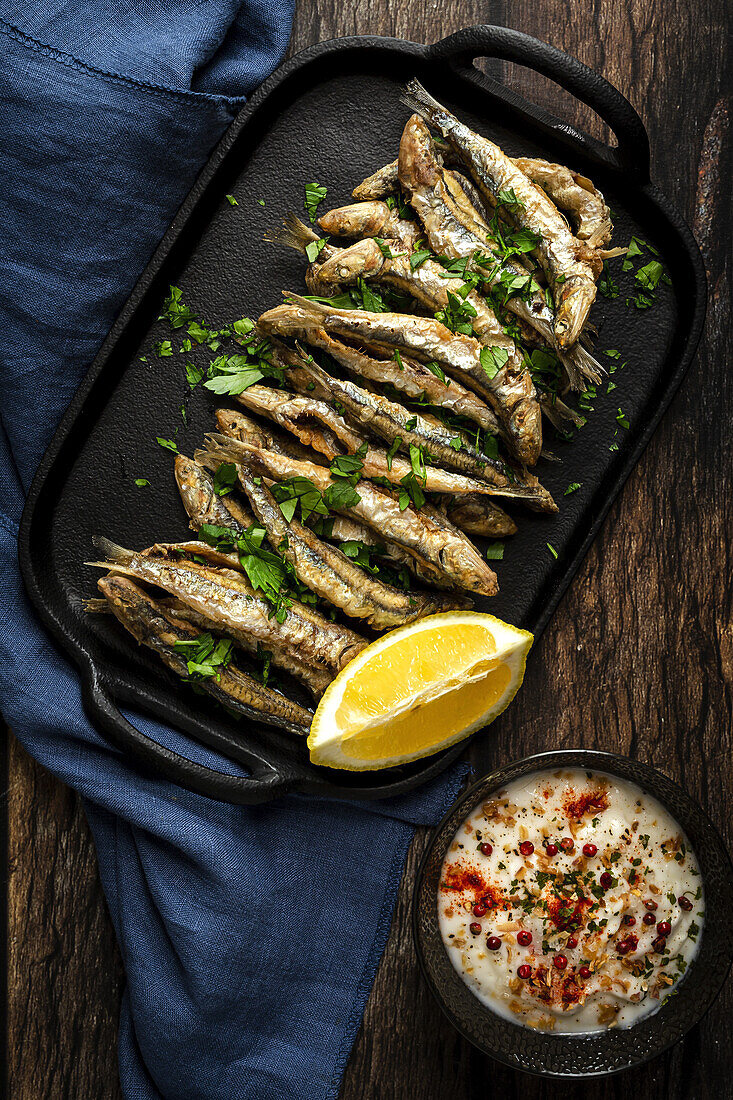 Top view of tasty fried anchovies with juicy lemon piece and chopped parsley on tray against savory sauce