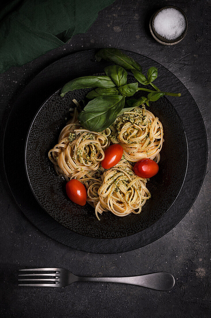 Top view of palatable Italian tagliatelle pasta with pesto sauce garnished with fresh red tomatoes and basil leaves served on black plate