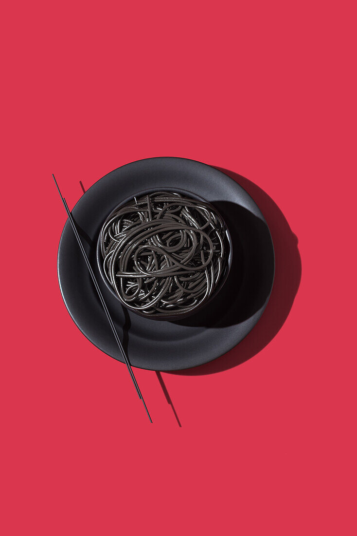 Draufsicht auf schwarze Spaghetti in einer Schüssel mit Stäbchen auf schwarzem und rotem Hintergrund