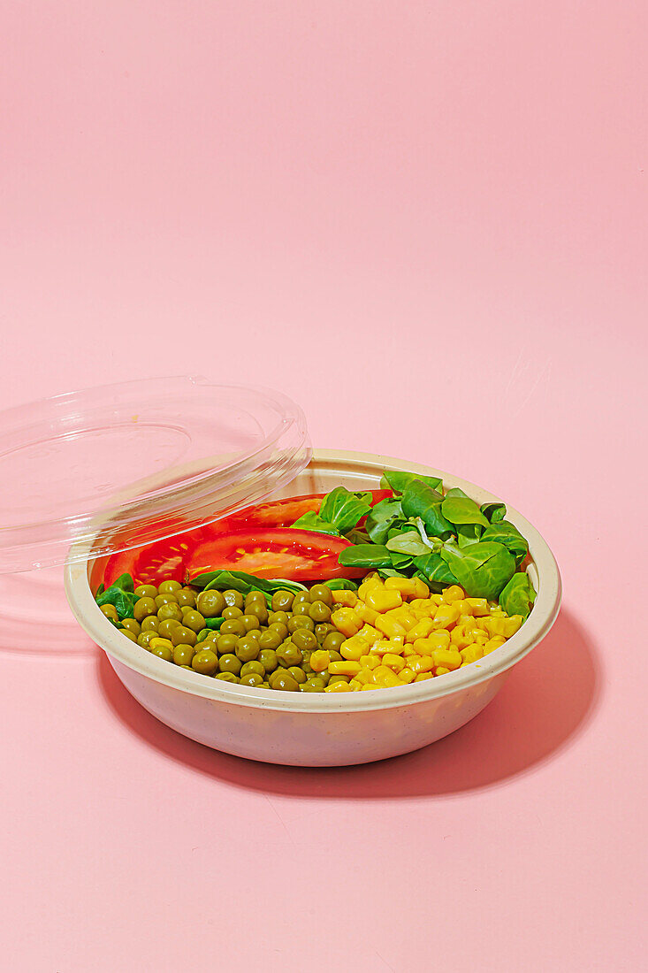 High angle of salad bowl with slices of tomato, spinach leaves, corn kernels and peas placed on pink surface