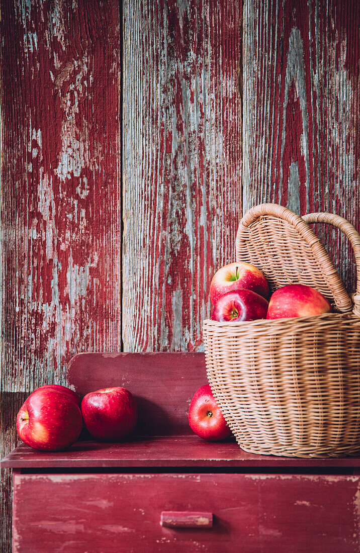 Wicker basket filled with fresh ripe red apples placed on shabby timber cupboard against wooden wall in countryside house during harvest season
