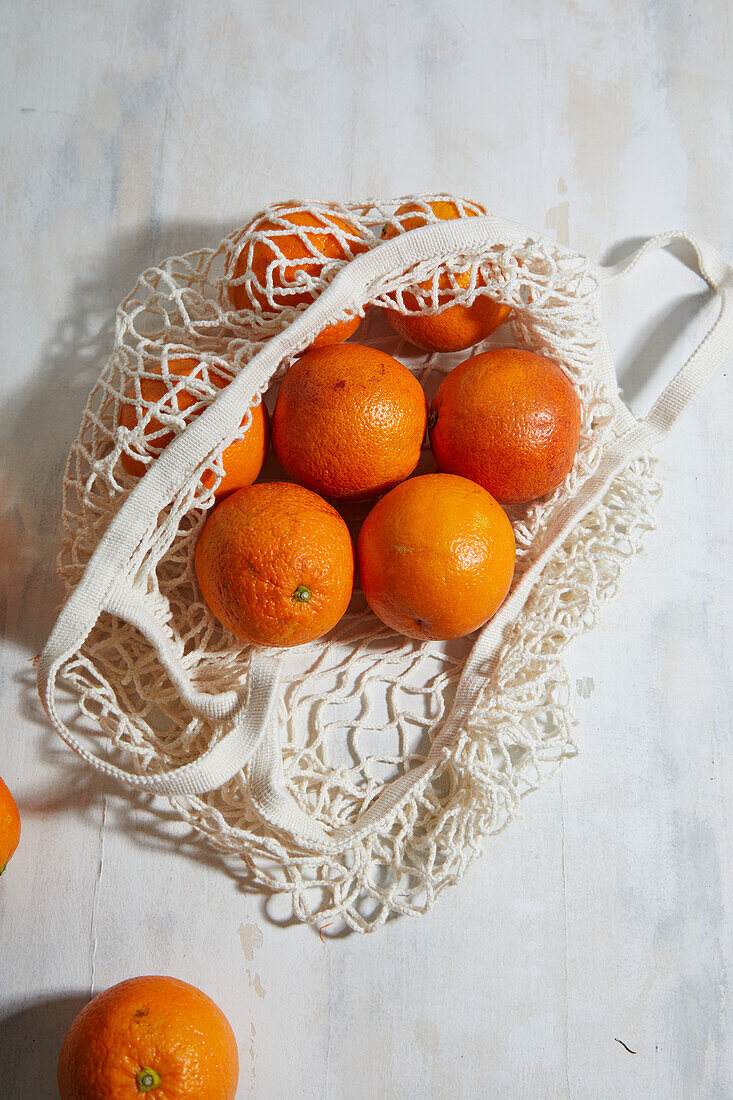 Draufsicht auf frische, ungeschälte blutige Orangen, die in einem wiederverwendbaren weißen Netzbeutel auf dem Tisch liegen