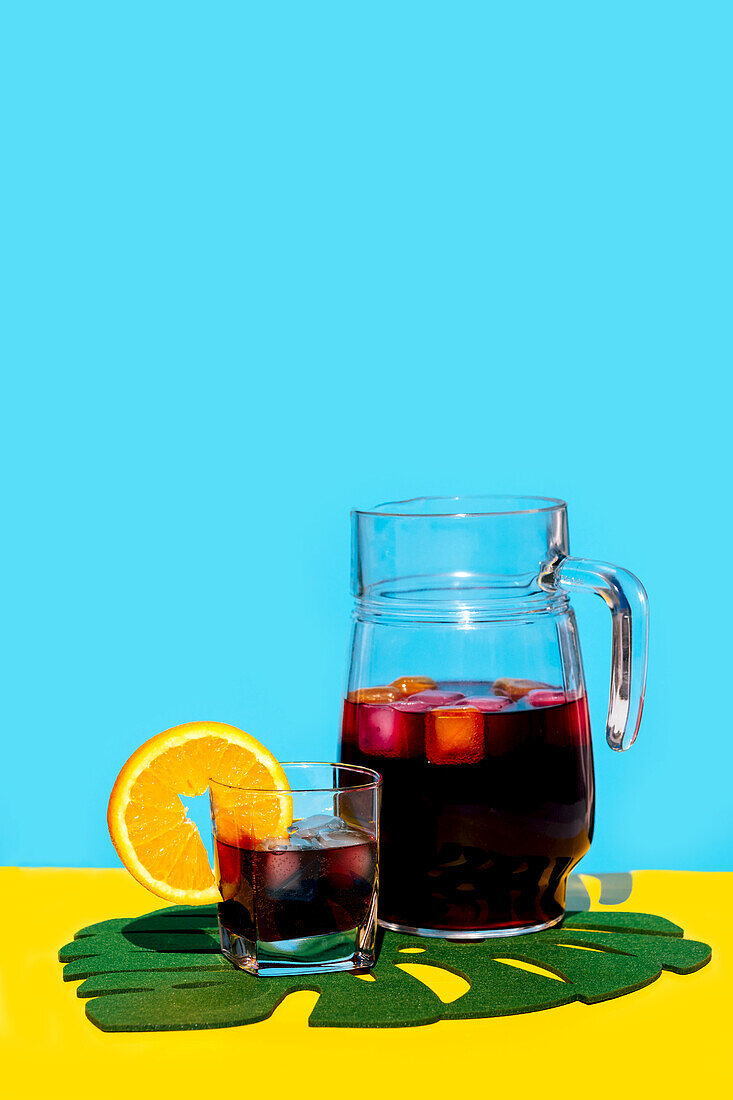 Kaltes Getränk mit Orangenscheibe im Glas und Krug auf Blattdekor über gelbem Hintergrund vor blauem Hintergrund im Studio
