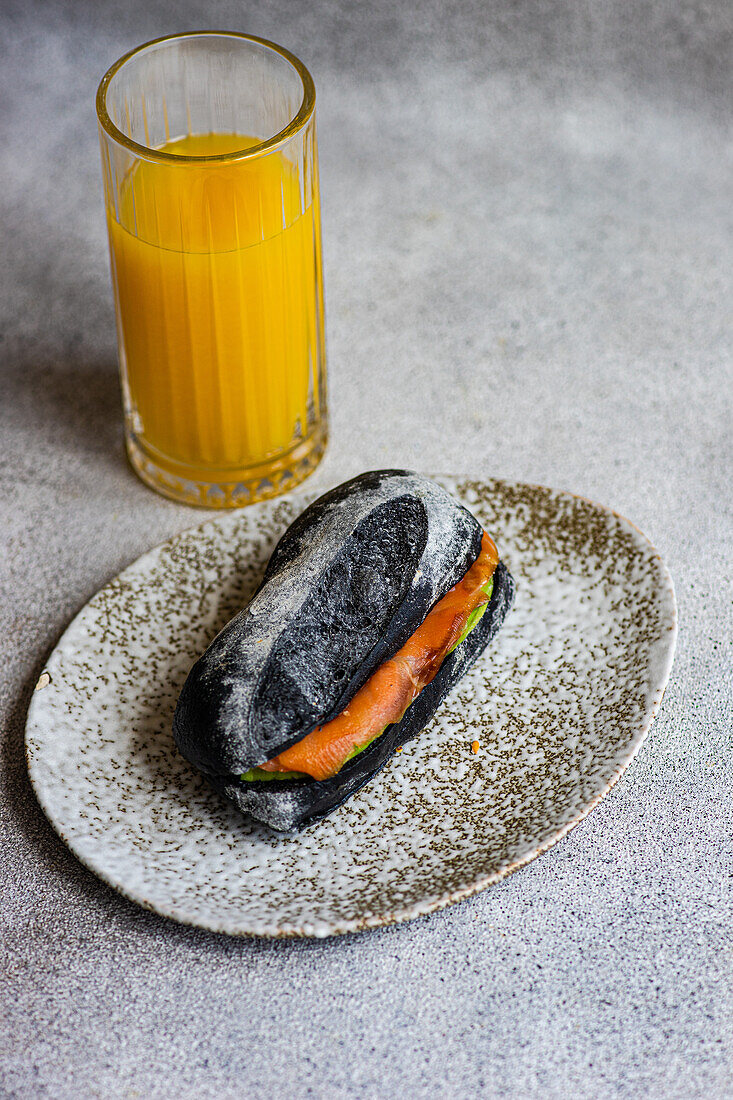 High angle of sandwich made of coal sour dough bread, salmon and avocado slices for breakfast served on marble plate near glass of juice against gray background